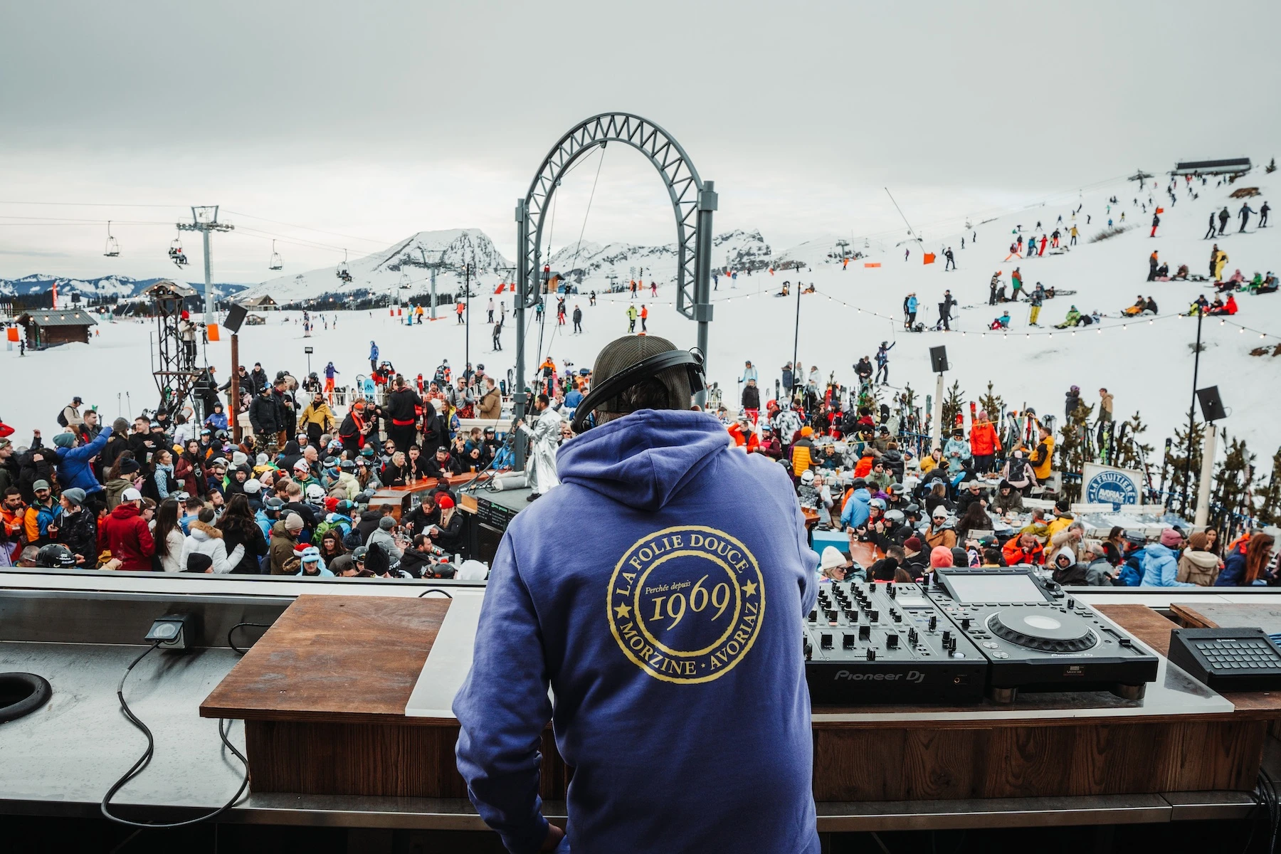 DJ wearing a blue hoodie with 'La Folie Douce 1969 Morzine Avoriaz' mixing music at a crowded outdoor snowy mountain ski resort event.