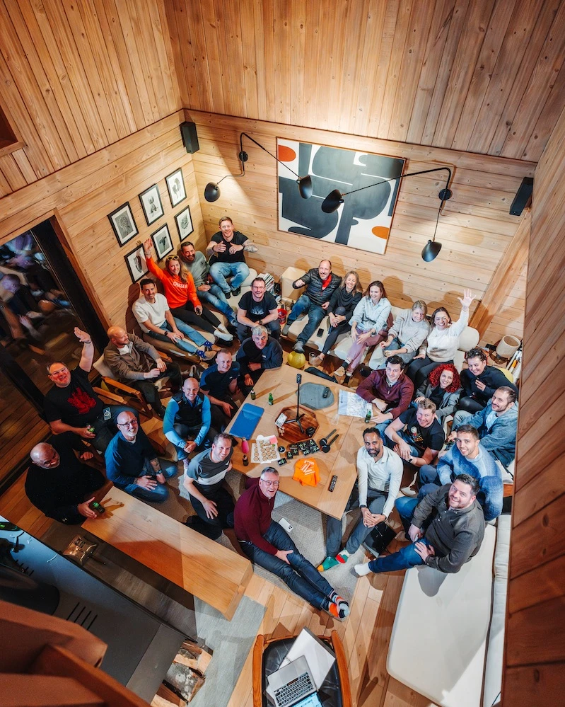Group of around 25 people sitting closely in a wooden-paneled room, smiling up at the camera with snacks and drinks on a central table.