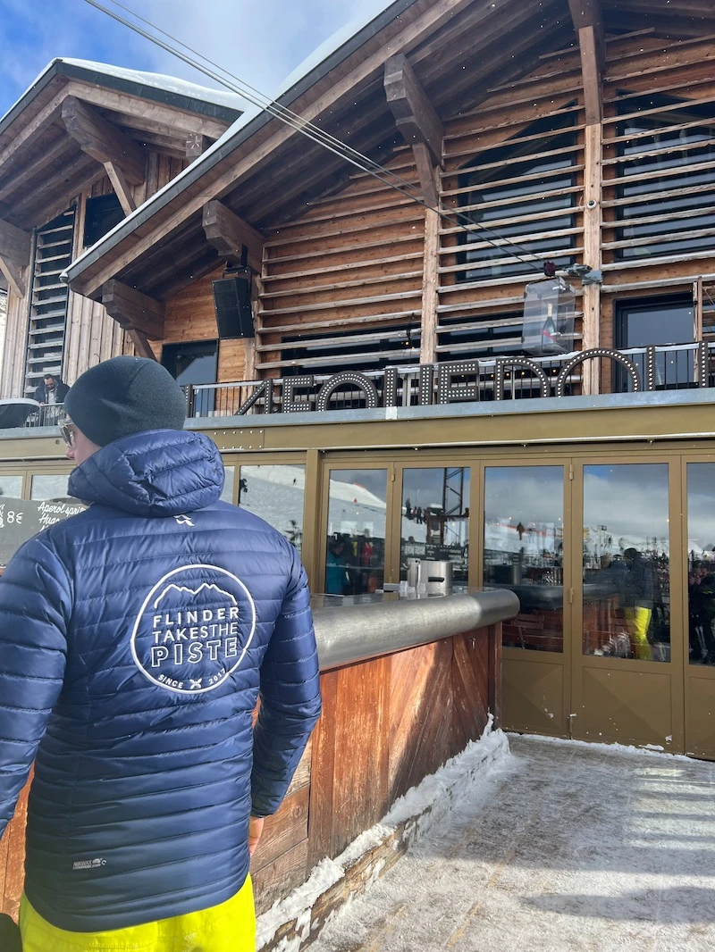Person in a blue jacket with fttp logo and black beanie standing near a wooden chalet