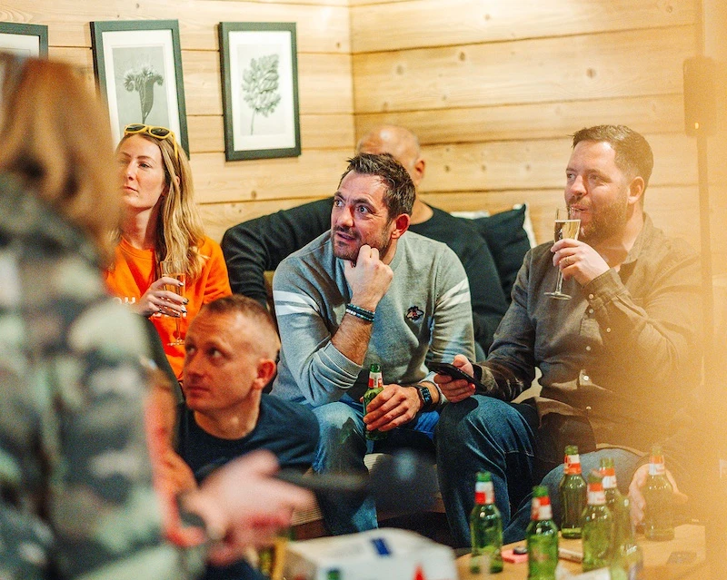 Group of five adults sitting closely in a cozy wooden room, socializing with drinks in hand and framed botanical prints on the wall.