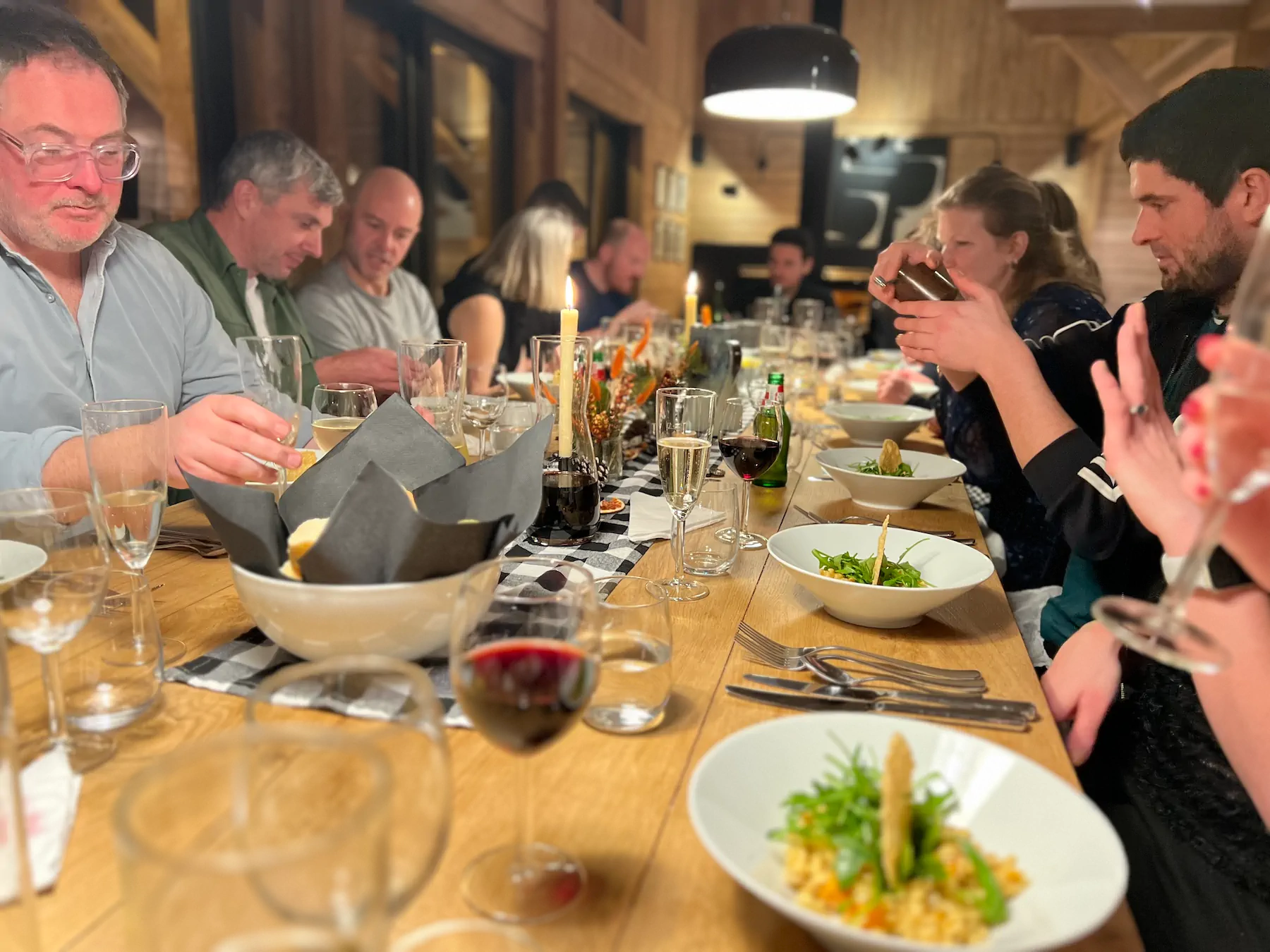 Group of people enjoying a meal at a long wooden dinner table with bowls of salad, glasses of wine, and lit candles.