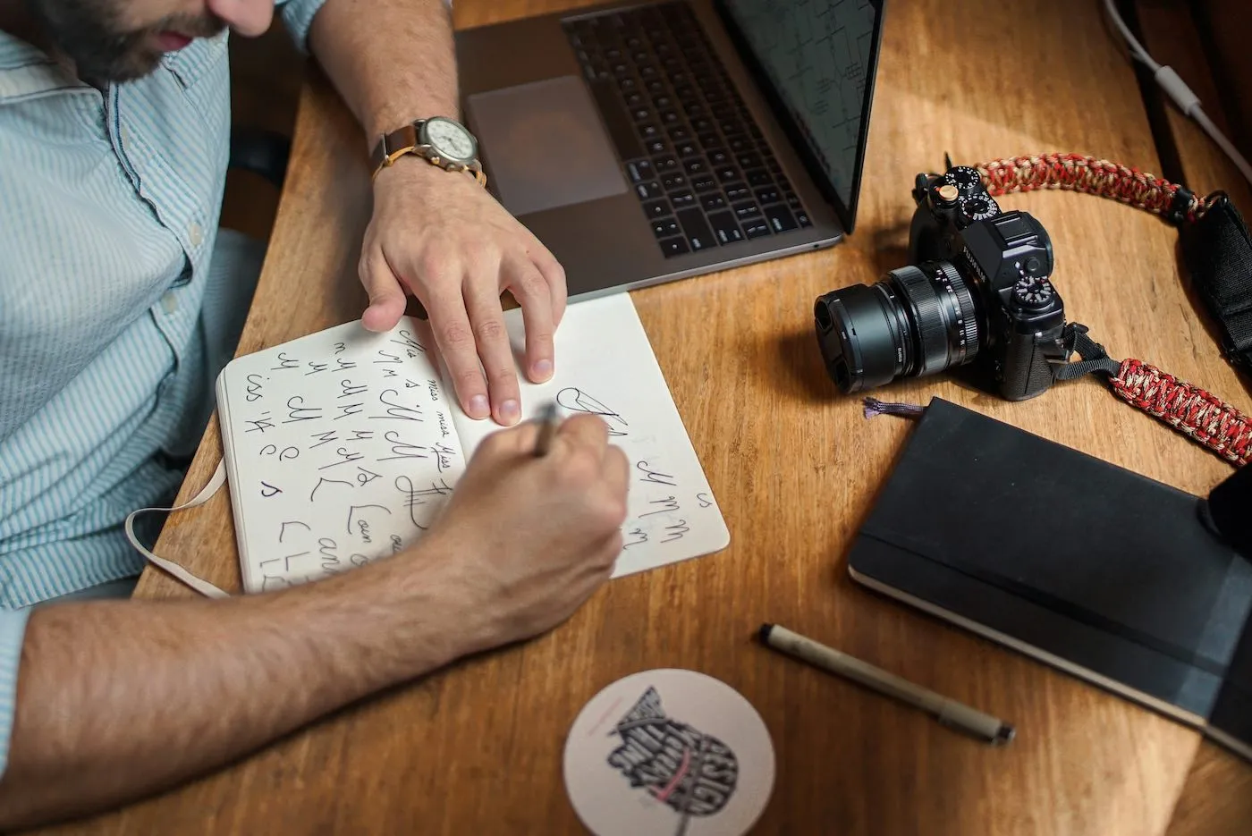 Person practicing calligraphy in a notebook at a wooden desk with a camera, closed notebook, and laptop nearby.