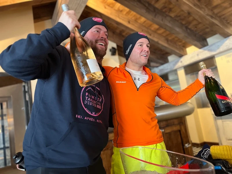 Two men wearing winter hats and sportswear smiling and holding large bottles of wine or champagne inside a wooden cabin.