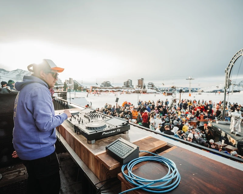 DJ wearing sunglasses and a cap performs on Pioneer DJ equipment at an outdoor snowy mountain event with a crowd gathered below.