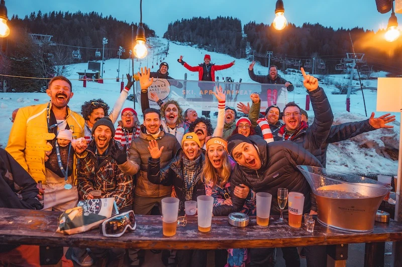 Group of cheerful people posing at a snowy ski resort at dusk with drinks on a wooden table and ski slopes in the background.