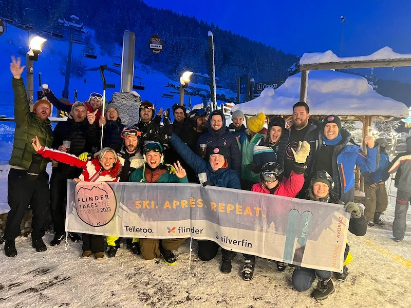 Group of people dressed in winter clothing holding a banner that reads 'Ski. Après. Repeat.' in a snowy outdoor ski resort at night.