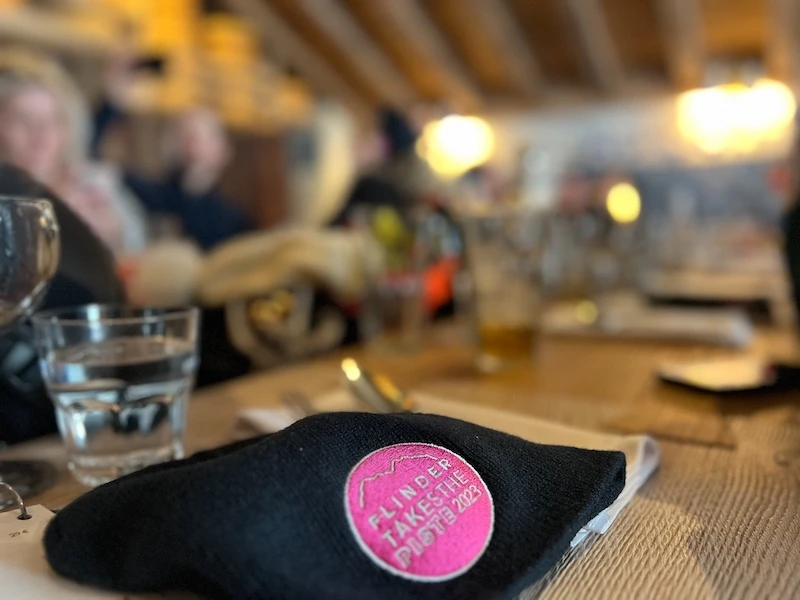 Close-up of a black cloth mask with a pink circular patch reading 'FLINDER TAKES THE PLATE 2023' on a wooden table in a blurred indoor setting.