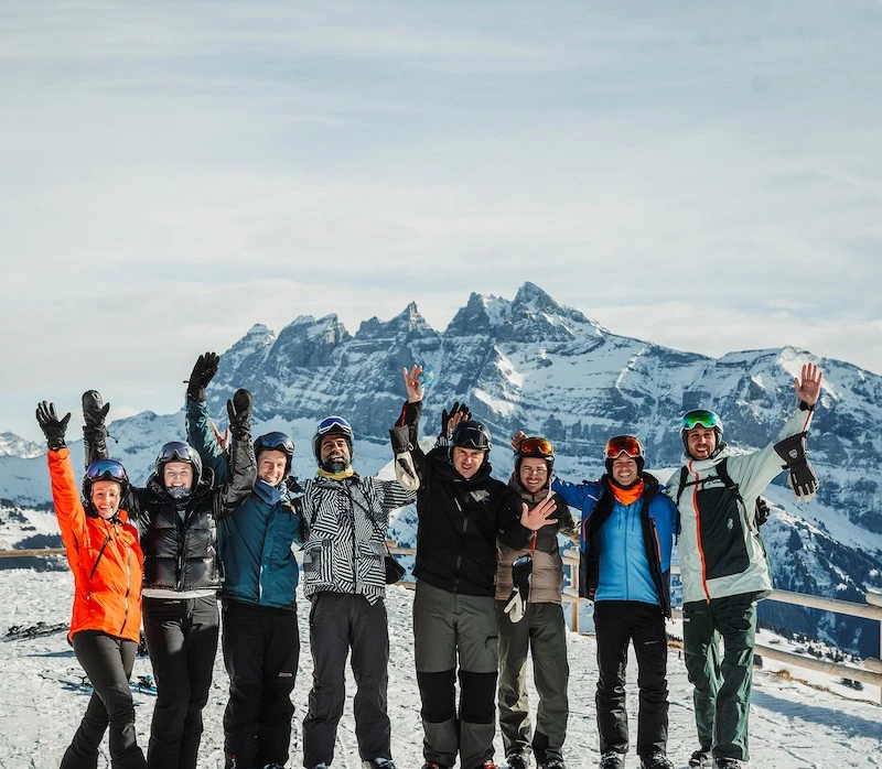 Group of eight people in ski gear standing on snow with arms raised and snow-covered mountains in the background.