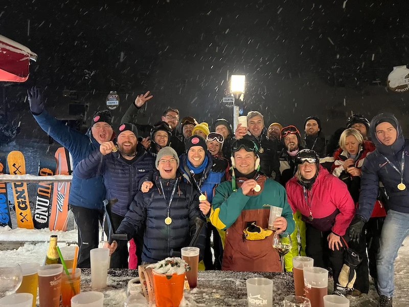 Group of people in winter clothing celebrating outdoors at night with snow falling, displaying medals and drinks on a snowy table.