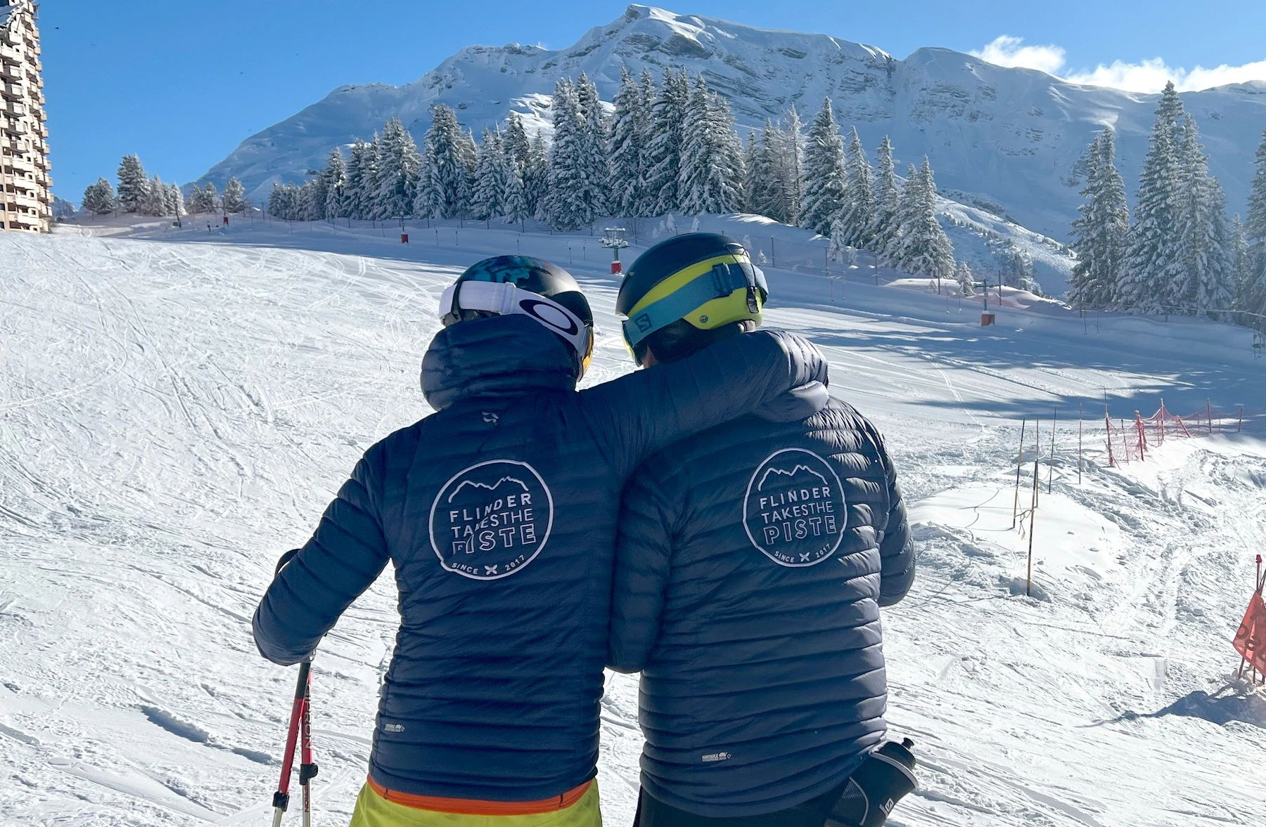 Two people wearing helmets and matching blue jackets with 'Flinder Takes the Piste' logo, embracing on a snowy ski slope with trees and mountains in the background.