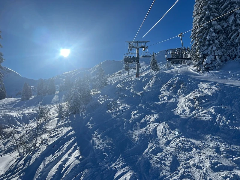 Ski lift carrying people up a snow-covered mountain slope under a bright sun and clear blue sky.