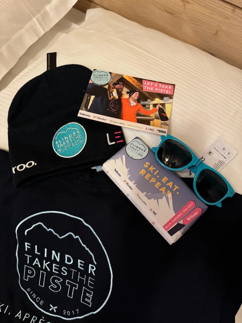 Collection of Flinder Takes the Piste 2024 merchandise including a black beanie, postcard, chocolate bar, blue sunglasses, and a black sweatshirt.