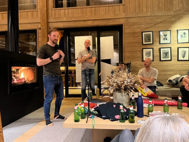 Two men holding microphones speaking in a cozy wooden room with a fireplace, surrounded by seated people and a table with drinks and snacks.
