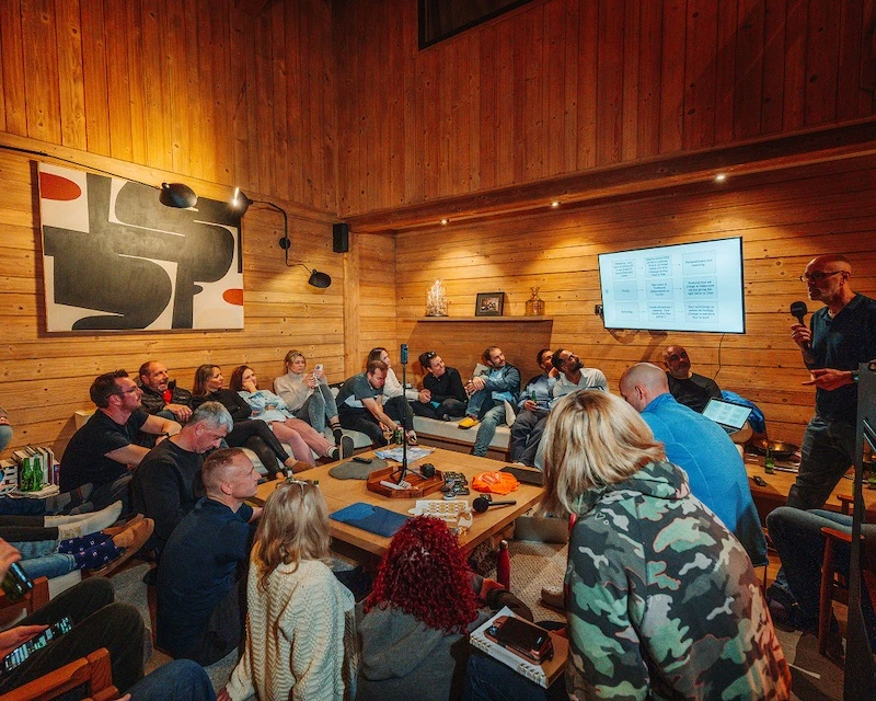 Group of people attentively watching a man giving a presentation with a microphone in a cozy wooden room.