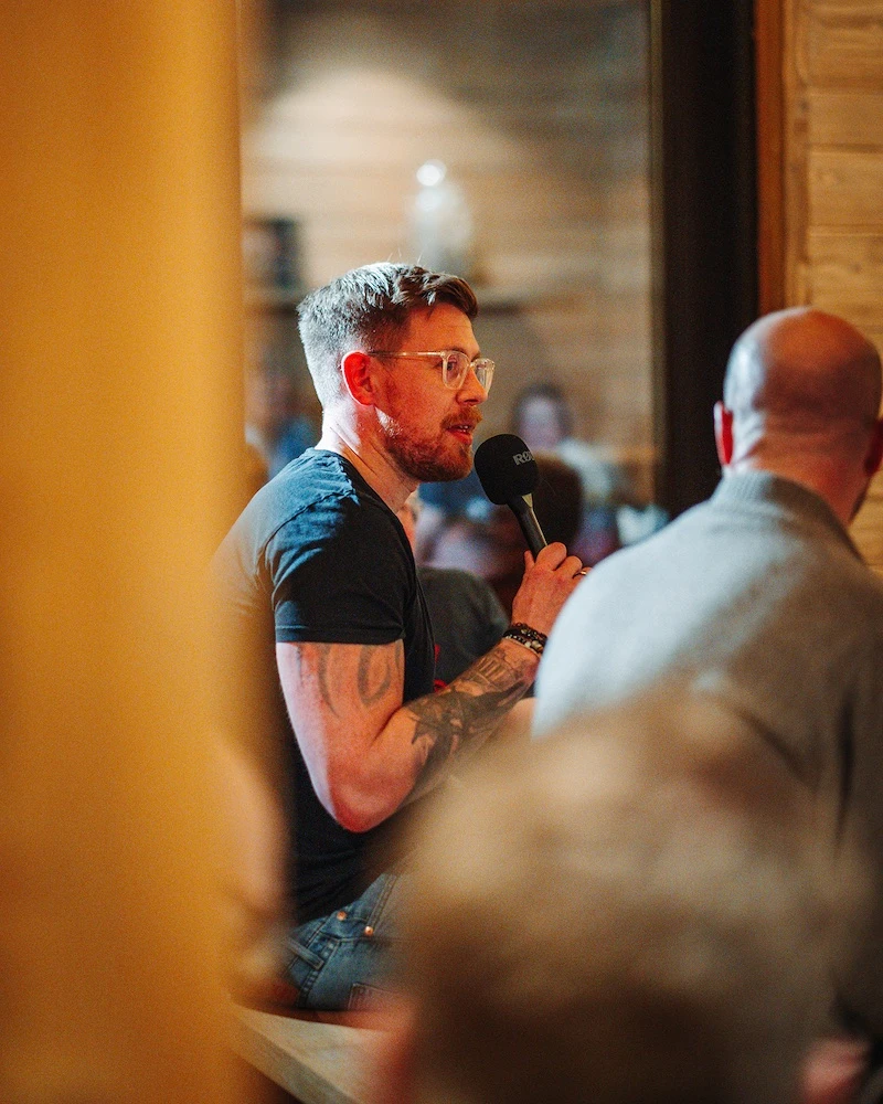 Man with glasses and tattoos speaking into a microphone in a cozy indoor setting.